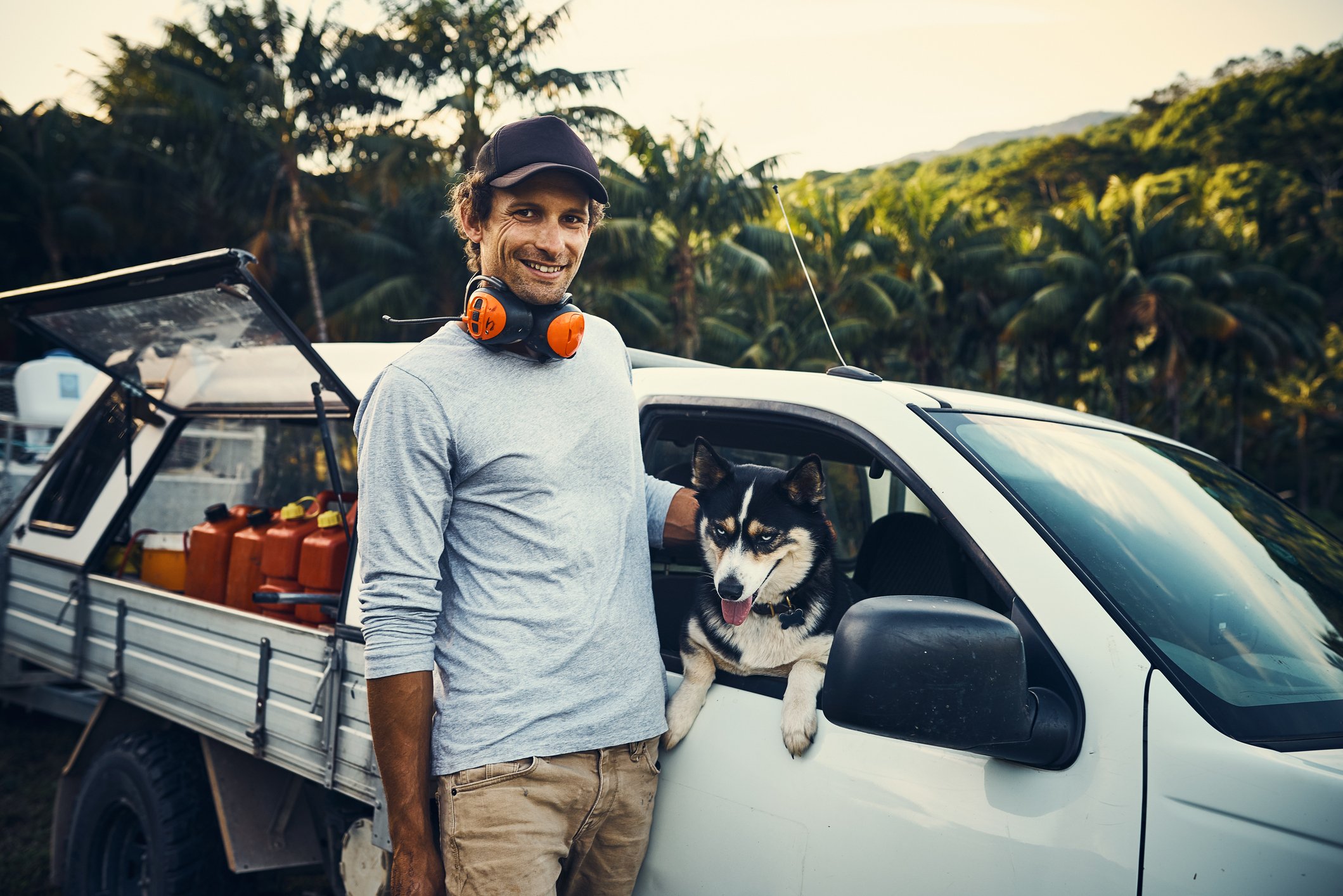 Man wearing work clothes and ear protectors petting his dog that is sitting inside a work truck