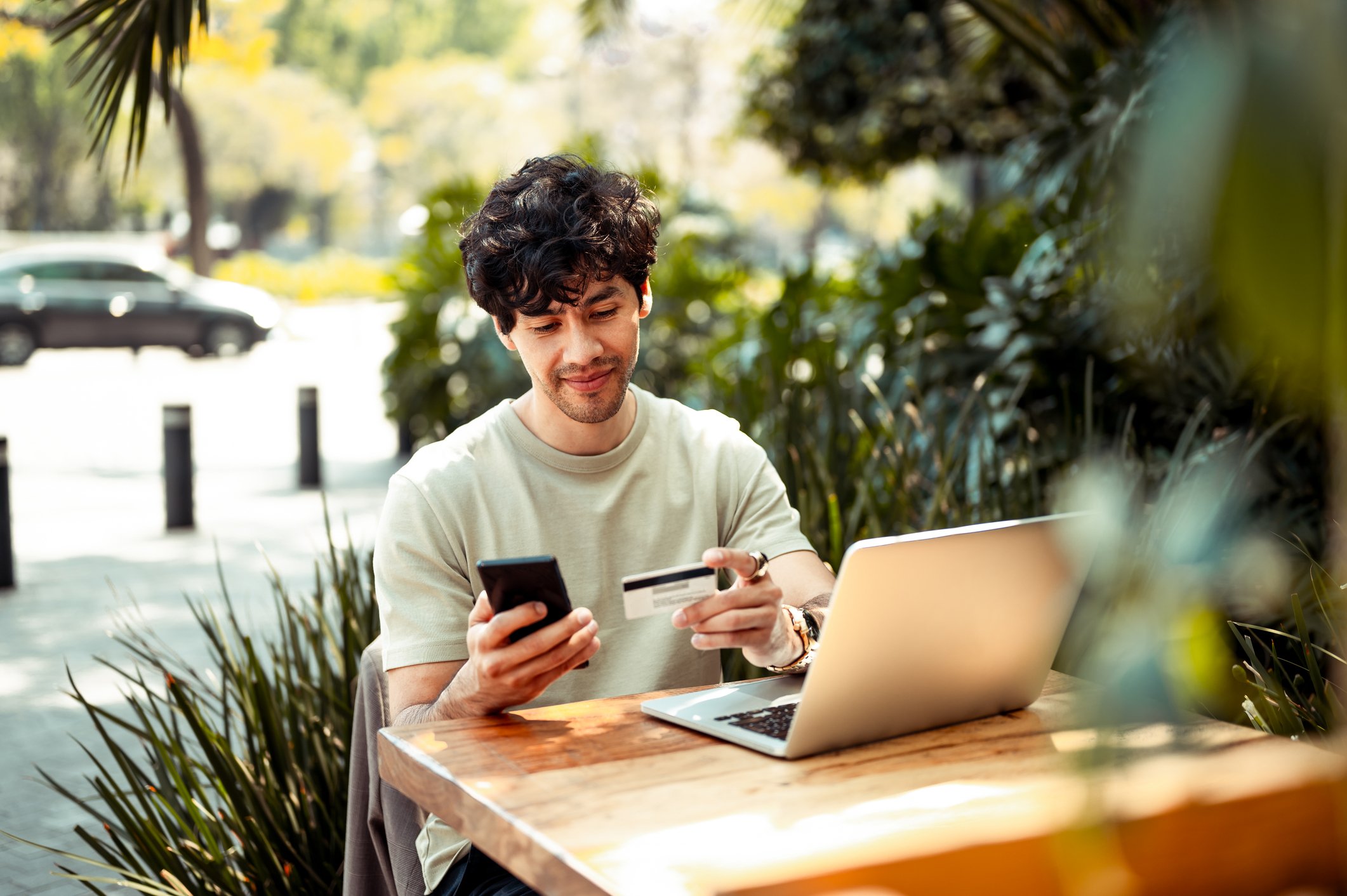 Young man using his credit card, smart phone and laptop outside at a table