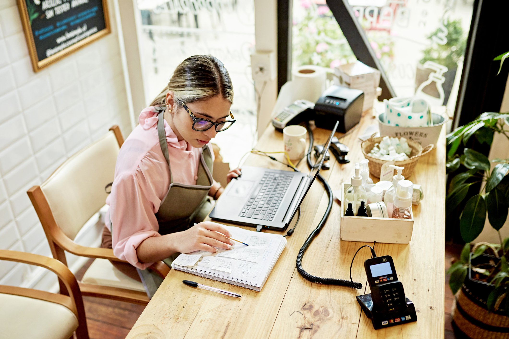 A woman wearing glasses sitting at a desk in a retail store looking at a notebook with her laptop in front of her